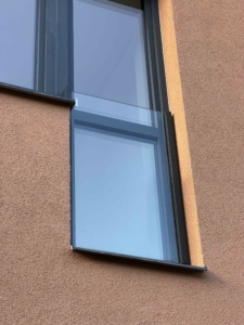 Detail moderního celoskleněného zábradlí na francouzském okně s bočním kotvením do hnědé fasády. Použito čiré bezpečnostní sklo 10mm a tmavé duralové konzoly. Realizace Studio Disa.