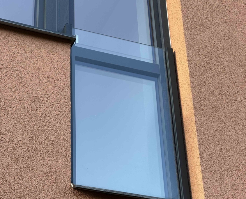 Detail moderního celoskleněného zábradlí na francouzském okně s bočním kotvením do hnědé fasády. Použito čiré bezpečnostní sklo 10mm a tmavé duralové konzoly. Realizace Studio Disa.